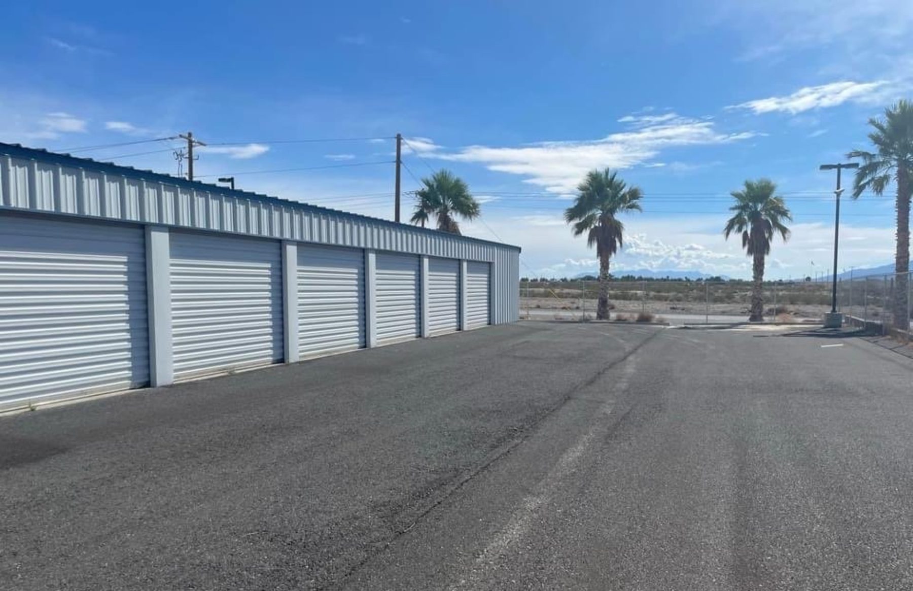 Paved drive aisle at PDMV1 Storage in Pahrump, lined with white roll-up doors, palm trees and open desert beyond the perimeter fence